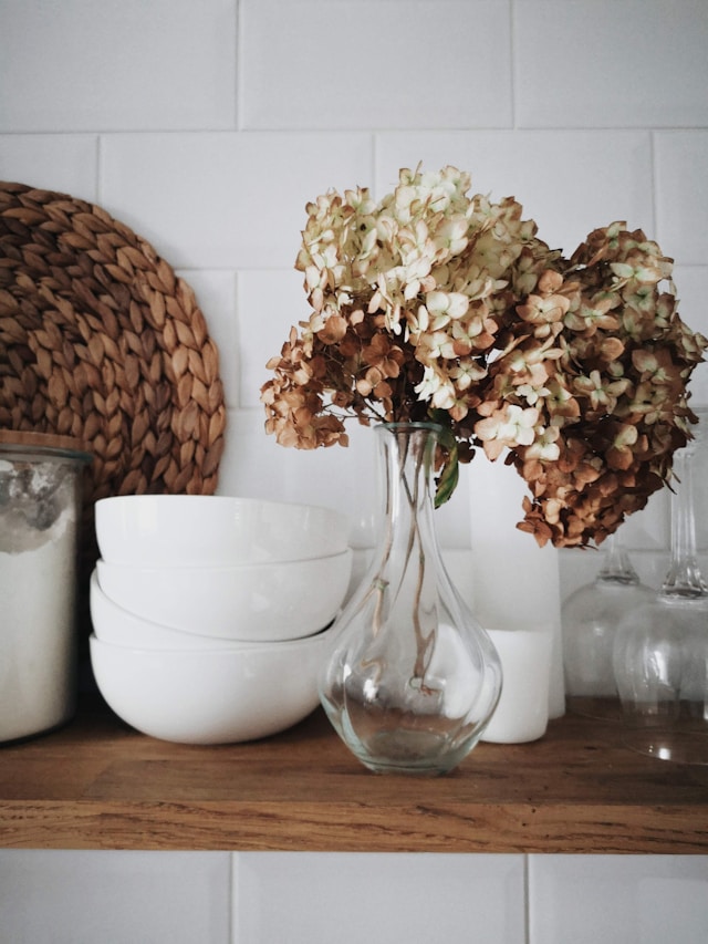 clear vase with fake beige and tan flowers on a kitchen shelf
